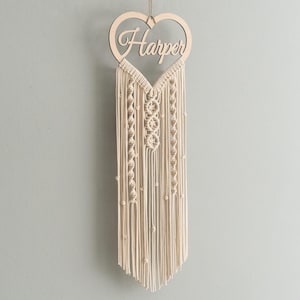 Puede incluir: Un tapiz de macramé blanco con un letrero de madera en forma de corazón que dice "Harper".