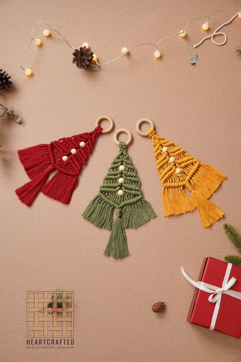 Puede incluir: Tres adornos de &aacute;rbol de Navidad de macram&eacute; hechos a mano en rojo, verde y amarillo con anillos de madera y cuentas blancas. Los adornos est&aacute;n colgados sobre un fondo marr&oacute;n con luces de cadena.
