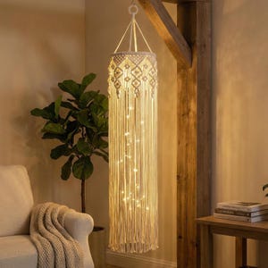May include: A cream-colored macrame pendant light fixture with a geometric design at the top and long, dangling fringe. The light is illuminated with small, warm white fairy lights, creating a cozy ambiance. The fixture hangs from a metal ring.