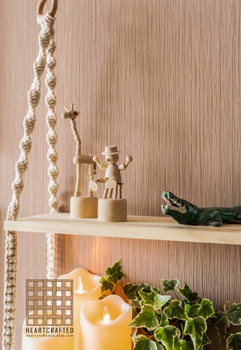 Może przedstawiać: Dwie drewniane p&oacute;łki z linowymi wieszakami, na kt&oacute;rych znajdują się drewniana żyrafa, drewniana marionetka, zielony ceramiczny aligator i doniczka z zielonymi liśćmi. P&oacute;łki znajdują się na tle imitującym drewno z trzema białymi świecami.