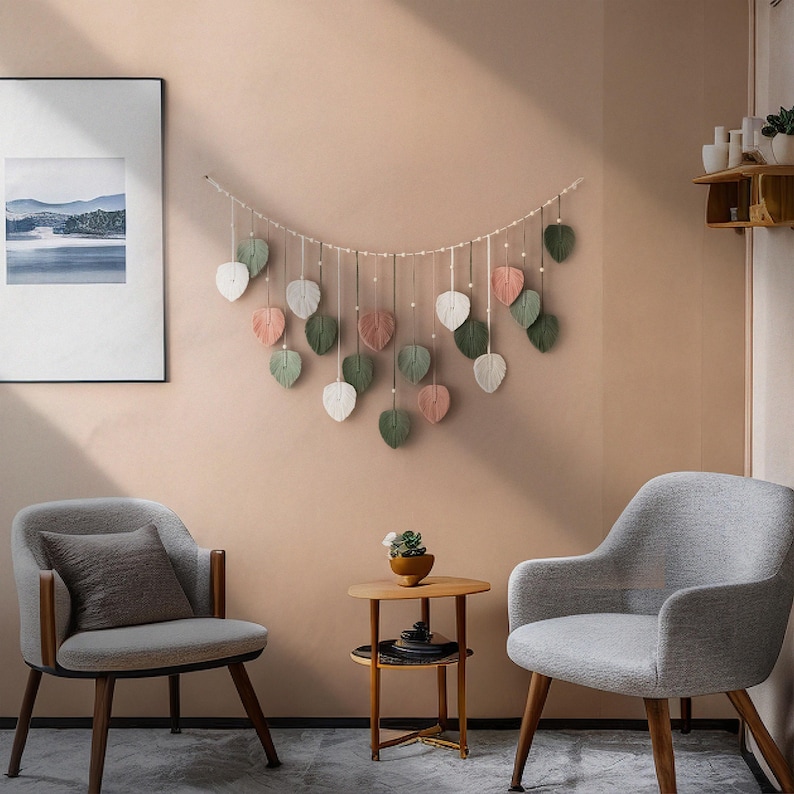 May include: A wall hanging with a string of leaves in shades of green, pink, and white. The leaves are made of yarn and are hanging from a string. The wall hanging is in front of a light brown wall. Two gray armchairs and a small wooden table are in the foreground.