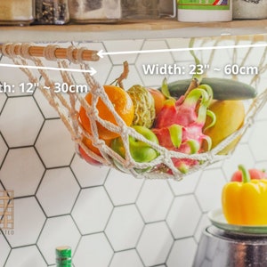 May include: White cotton rope hammock hanging from a wooden dowel rod, holding a variety of fruits and vegetables, including oranges, limes, dragon fruit, and cucumbers. The hammock is 23 inches wide and 12 inches deep.