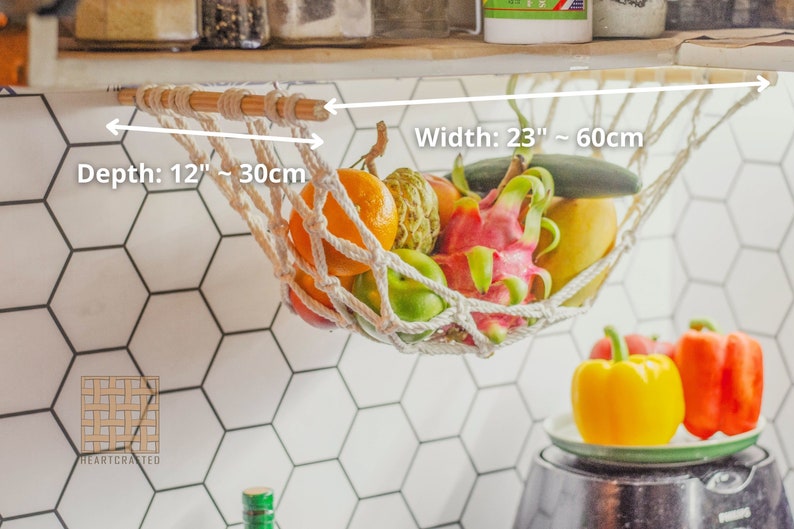 May include: White cotton rope hammock hanging from a wooden dowel rod, holding a variety of fruits and vegetables, including oranges, limes, dragon fruit, and cucumbers. The hammock is 23 inches wide and 12 inches deep.