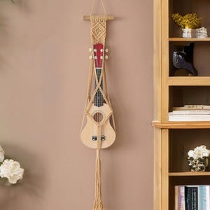 May include: A natural wood ukulele with a red headstock is suspended in a macrame plant hanger. The beige macrame has a geometric design at the top and a long tassel at the bottom. The ukulele is hanging on a light brown wall.