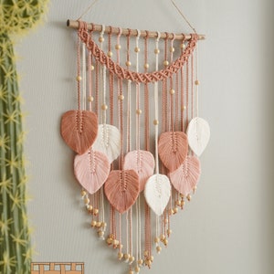 Op de afbeelding: Een macram&eacute; wandhanger met een houten lat aan de bovenkant. De hanger heeft een geweven ontwerp met roze en wit garen en houten kralen. Er hangen verschillende bladeren van hetzelfde garen aan de onderkant.
