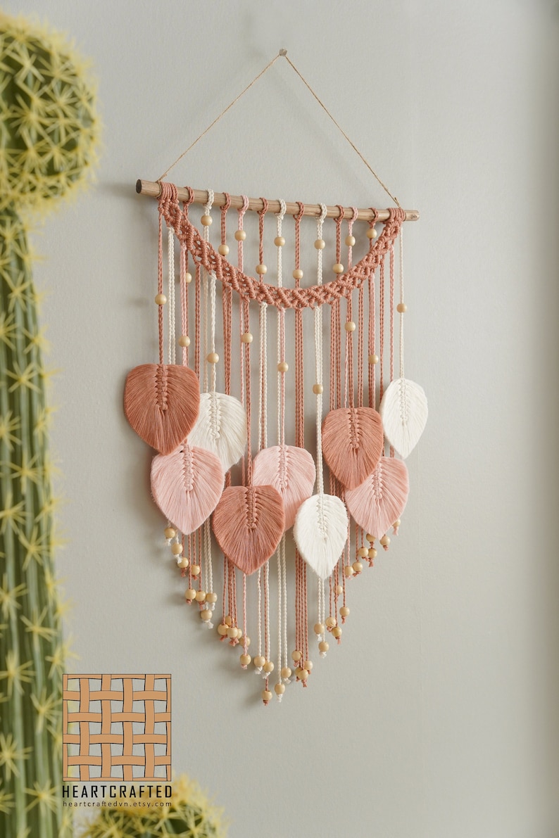 Op de afbeelding: Een macram&eacute; wandhanger met een houten lat aan de bovenkant. De hanger heeft een geweven ontwerp met roze en wit garen en houten kralen. Er hangen verschillende bladeren van hetzelfde garen aan de onderkant.