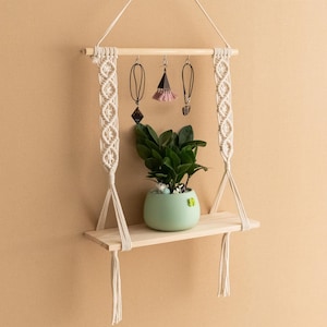 May include: A macrame hanging shelf with a wooden shelf and tassels. The shelf is decorated with jewellery and a small potted plant.