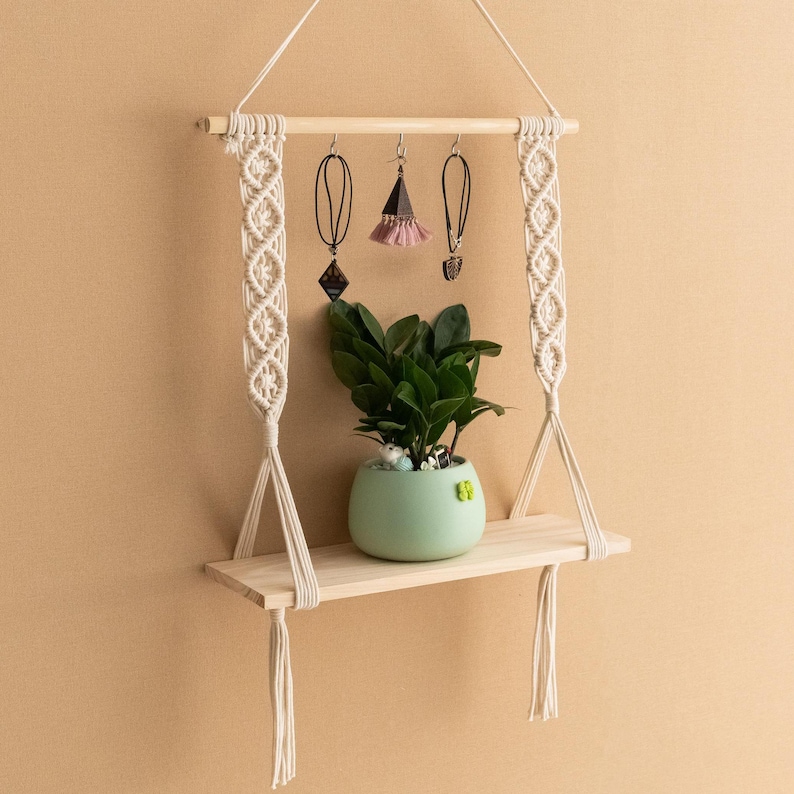 May include: A macrame hanging shelf with a wooden shelf and tassels. The shelf is decorated with jewellery and a small potted plant.