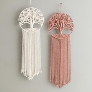 May include: Two macrame wall hangings, one cream and one dusty rose, each featuring a tree of life design within a circular frame. Long, fringed tassels hang below the tree, adding texture and visual interest. These decorative pieces are perfect for adding a touch of bohemian style to any room.