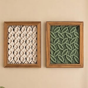 Pode incluir: Duas tapeçarias de parede em macramé emolduradas. Uma apresenta um padrão tecido creme, e a outra tem um design de folhas verdes. Ambas estão emolduradas em madeira, oferecendo uma estética natural e boêmia. Estas peças decorativas adicionam textura e interesse visual a qualquer espaço.