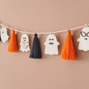 May include: A Halloween garland featuring white ghost cutouts and orange and black tassels. The ghosts have various facial expressions, including a heart, glasses, and a frown.