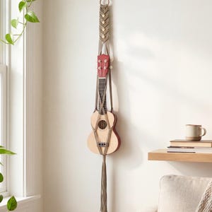 May include: A natural wood ukulele with a red headstock is suspended in a macrame plant hanger. The hanger is made of tan rope and features wooden beads. The ukulele is hanging on a white wall near a window and a small shelf.