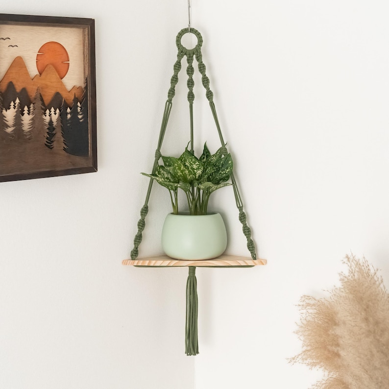 May include: A green macrame plant hanger with a wooden shelf and a green potted plant. The plant hanger is hanging from the ceiling and the shelf is attached to the hanger.