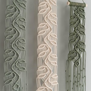May include: Three vertical macrame wall hangings suspended from light-colored wooden dowels. Each piece features a repeating leaf-like knot pattern in sage green, cream, and olive green cord, ending in long, loose fringe with small knotted accents.