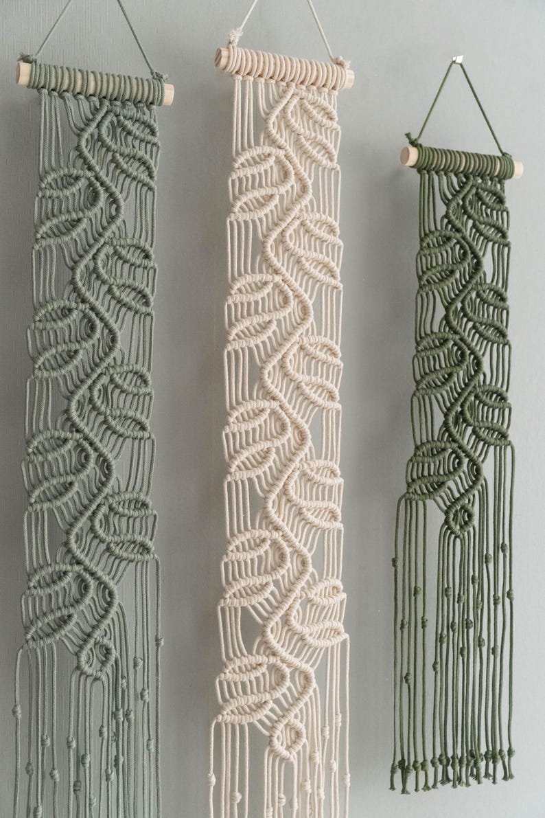 May include: Three vertical macrame wall hangings suspended from light-colored wooden dowels. Each piece features a repeating leaf-like knot pattern in sage green, cream, and olive green cord, ending in long, loose fringe with small knotted accents.