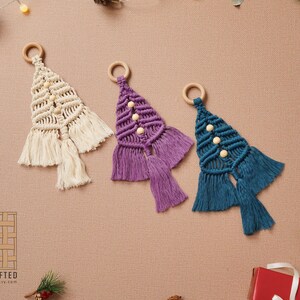 Puede incluir: Tres adornos de &aacute;rbol de Navidad de macram&eacute; hechos a mano en crema, morado y azul verdoso con cuentas de madera y flecos. Los adornos cuelgan de anillos de madera.