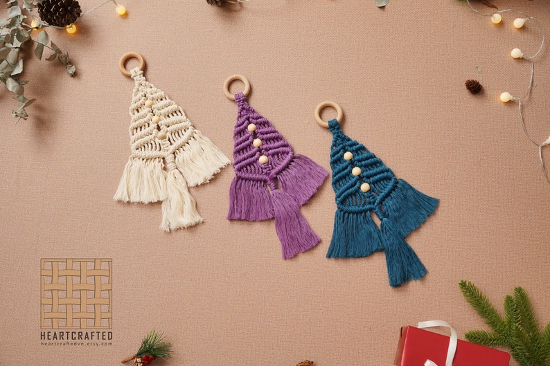 Puede incluir: Tres adornos de &aacute;rbol de Navidad de macram&eacute; hechos a mano en crema, morado y azul verdoso con cuentas de madera y flecos. Los adornos cuelgan de anillos de madera.