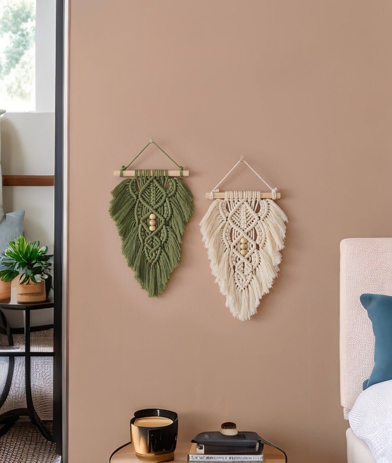 May include: Two macrame wall hangings, one green and one cream, with wooden dowels and beads. The hangings are displayed on a light brown wall.