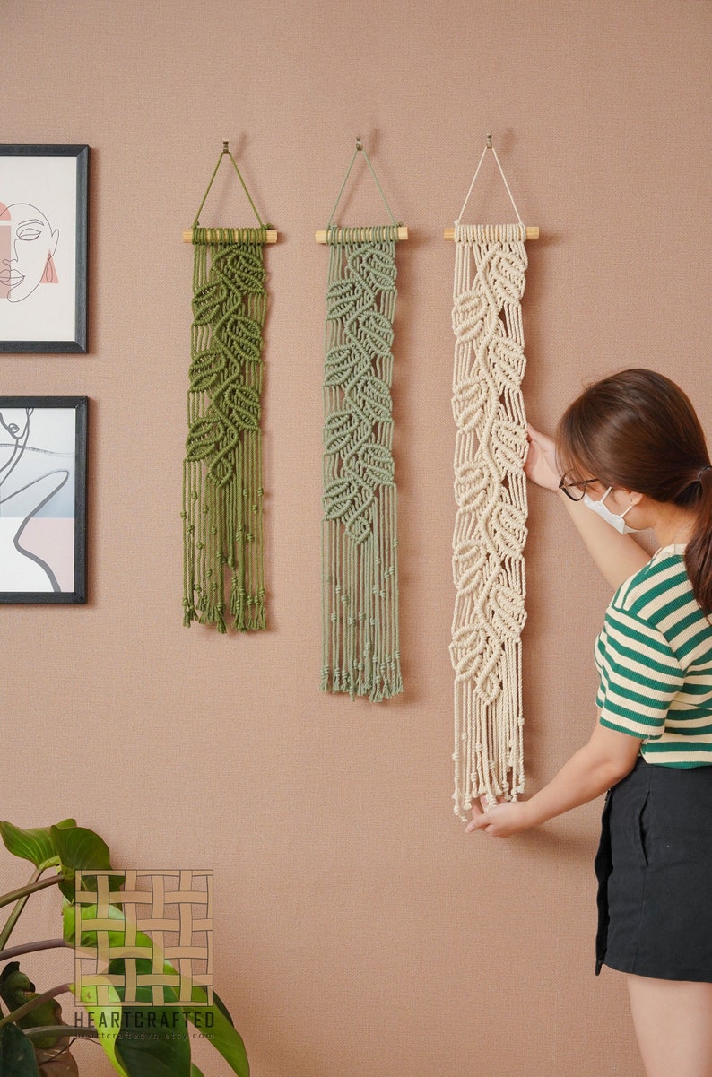 May include: Three macrame wall hangings with a leaf design. The hangings are in shades of green and white. The hangings are hung on a light brown wall.