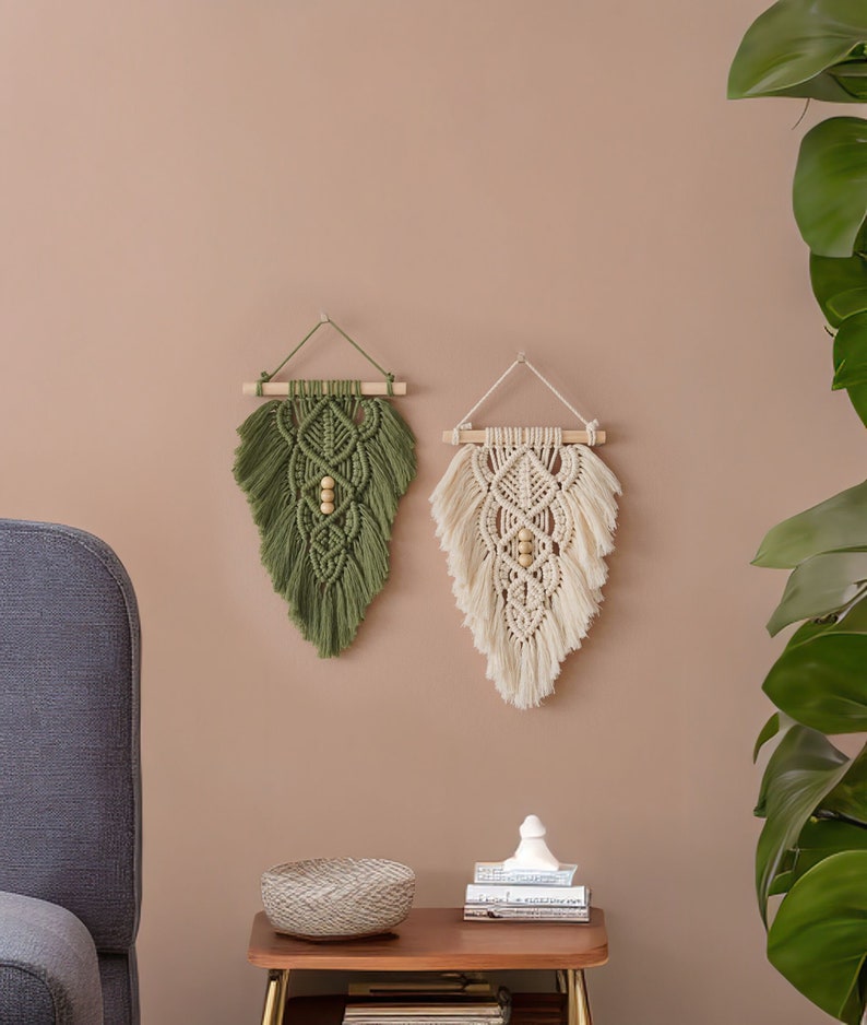 May include: Two macrame wall hangings, one in green and one in cream, with wooden dowels and wooden beads. The hangings are displayed on a light brown wall.