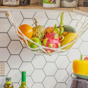 May include: A white hammock hanging from a kitchen cabinet, filled with a variety of fresh fruits, including a dragon fruit, a pineapple, a mango, a lime, and a starfruit. The hammock is hanging over a white hexagonal tile backsplash.