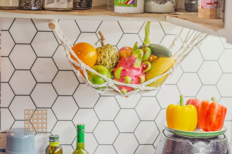 May include: A white hammock hanging from a kitchen cabinet, filled with a variety of fresh fruits, including a dragon fruit, a pineapple, a mango, a lime, and a starfruit. The hammock is hanging over a white hexagonal tile backsplash.