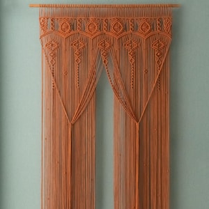 May include: An orange macrame wall hanging with a geometric design. The top features a series of repeating diamond shapes, and the lower portion has long, vertical strands. The piece is suspended from a wooden dowel.