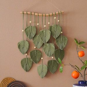 May include: A macramé wall hanging with olive green leaf shapes suspended from a natural wooden dowel. Each leaf is detailed with intricate knotting and accented with small wooden beads. The wall decor is a bohemian style.