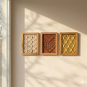 Pu&ograve; includere: Tre arazzi da parete in macram&egrave; incorniciati. Il primo presenta un design color crema, il secondo un design ruggine e il terzo un design giallo con perline bianche. Tutti sono in cornici di legno marrone chiaro, esposti su una parete beige.