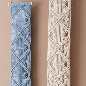 May include: Two macrame wall hangings, one in light blue and one in white, with a diamond pattern. Both hangings are made with cotton cord and have wooden dowels at the top.