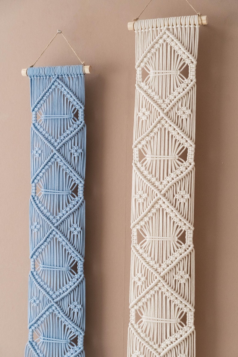 May include: Two macrame wall hangings, one in light blue and one in white, with a diamond pattern. Both hangings are made with cotton cord and have wooden dowels at the top.