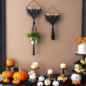 Pu&ograve; includere: Due arazzi da parete in macram&eacute; nero a forma di pipistrelli, uno con una pianta sospesa, con una variet&agrave; di zucche bianche e arancioni, candele e altre decorazioni di Halloween su una superficie nera.