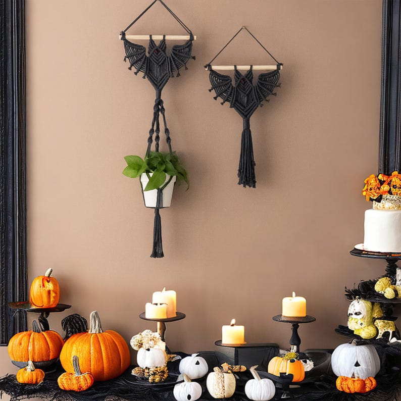 Pu&ograve; includere: Due arazzi da parete in macram&eacute; nero a forma di pipistrelli, uno con una pianta sospesa, con una variet&agrave; di zucche bianche e arancioni, candele e altre decorazioni di Halloween su una superficie nera.