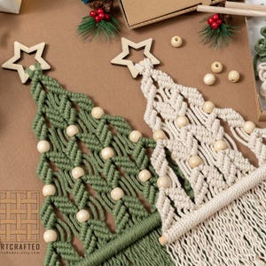 Puede incluir: Dos árboles de Navidad de macramé, uno verde y otro blanquecino, cada uno adornado con estrellas de madera y cuentas esféricas. Los árboles se exhiben sobre una superficie marrón, sugiriendo un tema festivo navideño.