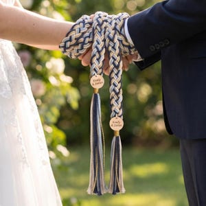 Puede incluir: Un cordón de ceremonia con una trenza gruesa de varios hilos en azul marino, azul claro y crema. El cordón termina en dos etiquetas circulares de madera grabadas con el texto Emily & James, con borlas largas y sueltas a juego.