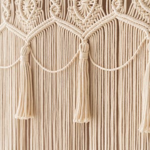 May include: Cream-colored macrame wall hanging with intricate woven patterns and decorative tassels. The design features a series of repeating geometric shapes and vertical strands, creating a textured, bohemian aesthetic. This handcrafted piece adds a touch of natural elegance to any space.