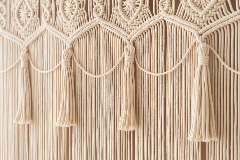 May include: Cream-colored macrame wall hanging with intricate woven patterns and decorative tassels. The design features a series of repeating geometric shapes and vertical strands, creating a textured, bohemian aesthetic. This handcrafted piece adds a touch of natural elegance to any space.