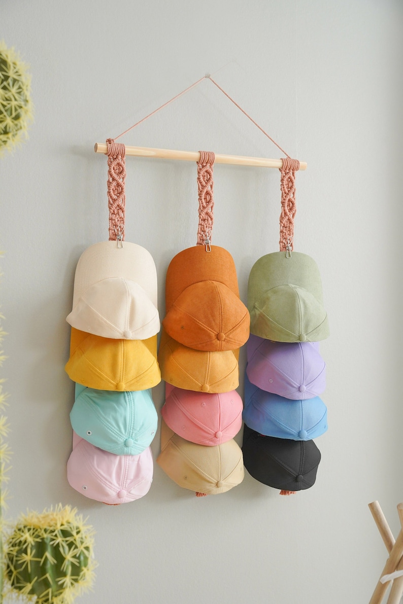 May include: A wooden hat rack with macrame hangers displays a collection of colorful baseball caps. The caps are in shades of cream, yellow, teal, pink, orange, peach, olive green, lavender, blue, and black. The rack is mounted on a light gray wall.