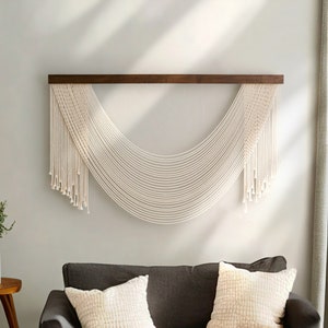 May include: A white macrame wall hanging with a wooden dowel rod, hanging on a light gray wall. The macrame is in the shape of a half-circle with long strands hanging down.