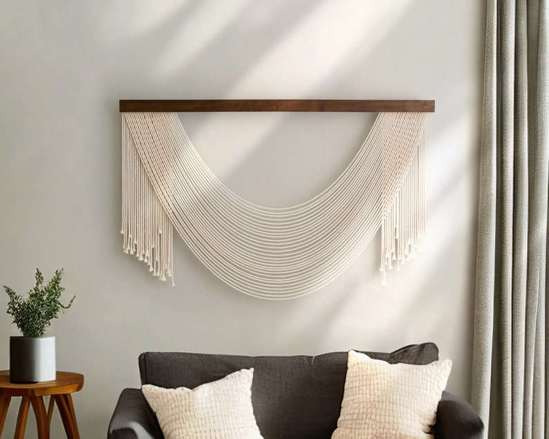 May include: A white macrame wall hanging with a wooden dowel rod, hanging on a light gray wall. The macrame is in the shape of a half-circle with long strands hanging down.