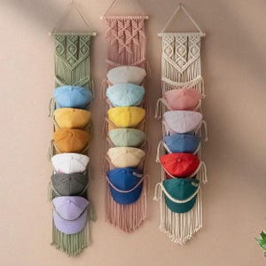 Puede incluir: Tres tapices de macramé verticales en verde salvia, rosa empolvado y crema, cada uno con intrincados patrones de nudos y múltiples bucles horizontales diseñados para sostener y exhibir una colección de gorras de béisbol de colores sólidos.