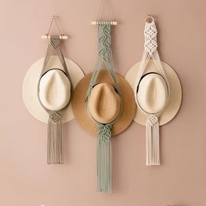 Puede incluir: Tres colgadores de sombreros de macramé en gris, verde salvia y blanco, cada uno sosteniendo un sombrero de paja. Los colgadores están suspendidos de la pared, mostrando un estilo bohemio. Los sombreros tienen una banda negra y un ala de color claro.