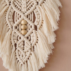 May include: A beige macrame wall hanging with wooden beads and fringe. The wall hanging is in a geometric pattern.