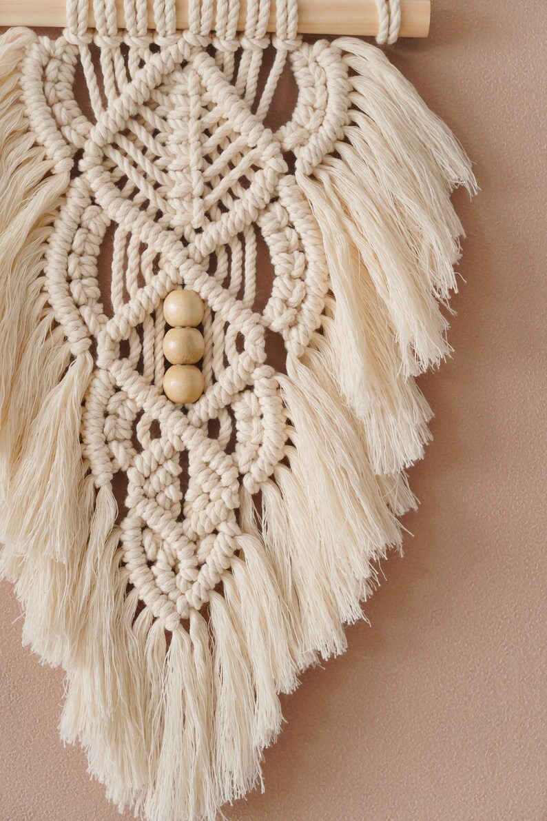 May include: A beige macrame wall hanging with wooden beads and fringe. The wall hanging is in a geometric pattern.