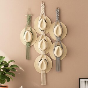 May include: Several straw hats are hung on a wall using macrame. The hats have a light-colored brim and a dark band. The macrame hangers are in white, grey, and green, providing a decorative element to the display. The wall is a neutral colour.