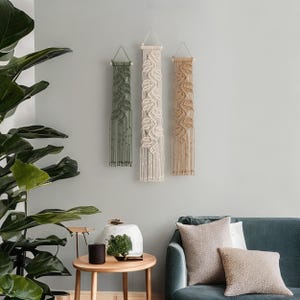 May include: Three vertical macrame wall hangings suspended from wooden dowels with string loops. The left hanging is olive green, the center is cream, and the right is tan. Each features intricate knotted leaf patterns and long fringe.