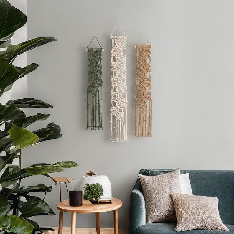 May include: Three vertical macrame wall hangings suspended from wooden dowels with string loops. The left hanging is olive green, the center is cream, and the right is tan. Each features intricate knotted leaf patterns and long fringe.