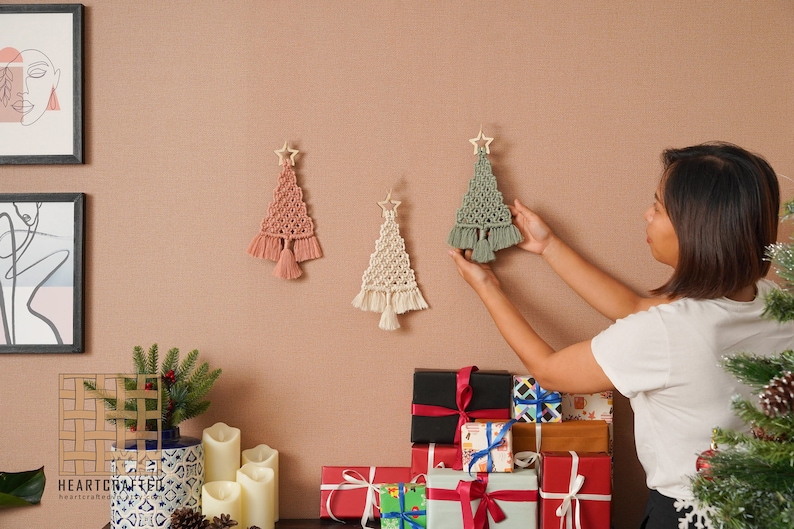 K&ouml;nnte beinhalten: Drei Makramee-Weihnachtsbaum-Wandbeh&auml;nge in Rosa, Wei&szlig; und Gr&uuml;n. Die Beh&auml;nge sind mit Holzsternen und Quasten verziert. Eine Frau h&auml;ngt einen der B&auml;ume an eine Wand.