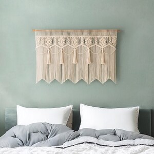 May include: Cream-colored macrame wall hanging with a geometric design and fringe. The textile is mounted on a wooden dowel and hangs above a bed with white pillows and a gray comforter. The wall is painted a muted green.