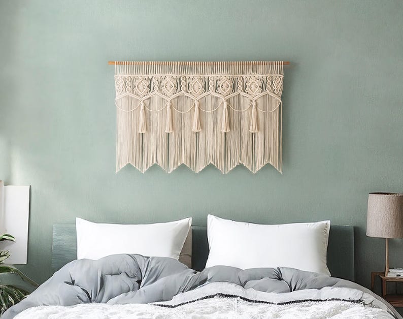 May include: Cream-colored macrame wall hanging with a geometric design and fringe. The textile is mounted on a wooden dowel and hangs above a bed with white pillows and a gray comforter. The wall is painted a muted green.